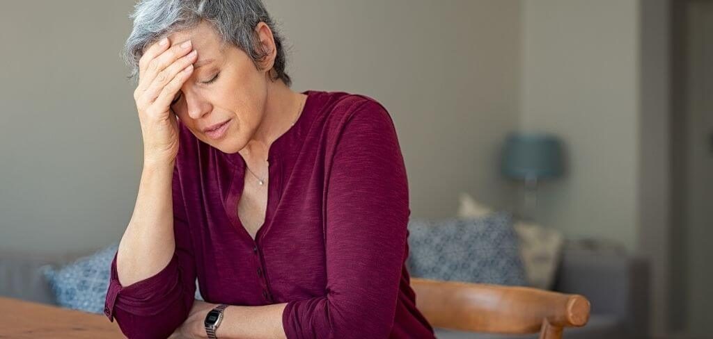 mujer-perfil-hormonal-beneficios-lab Mujer adulta con dolor de cabeza, sentada en su comedor con las manos en la cabeza.